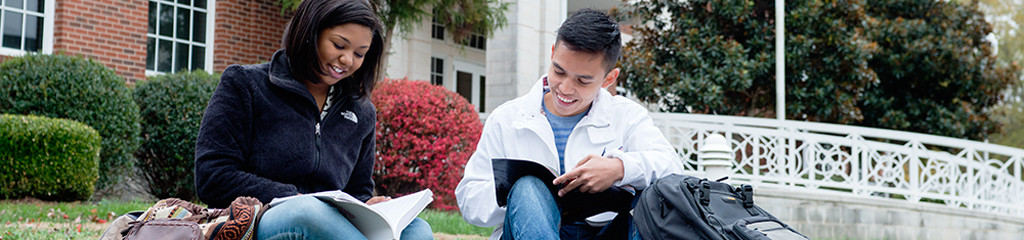 Male and female students sit outside of an academic building and look at their textbooks