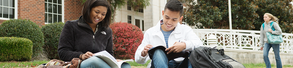 male and female student read textbooks on lawn in from of academic building