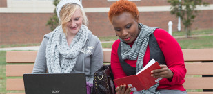 Two female students discuss a book on a bench outside on campus
